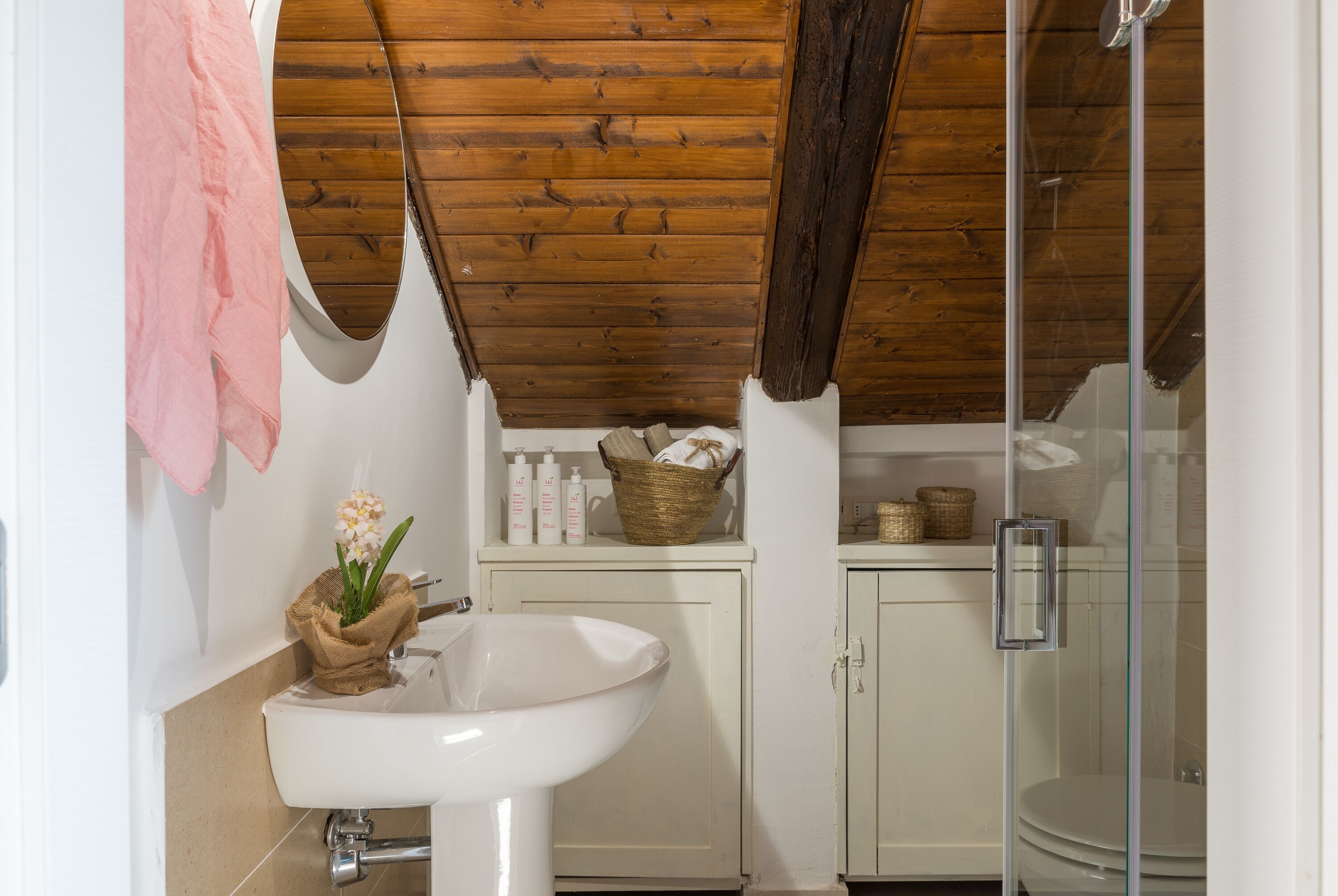 A bathroom with a wooden ceiling, a white sink, a glass shower enclosure, and a shelf with toiletries.