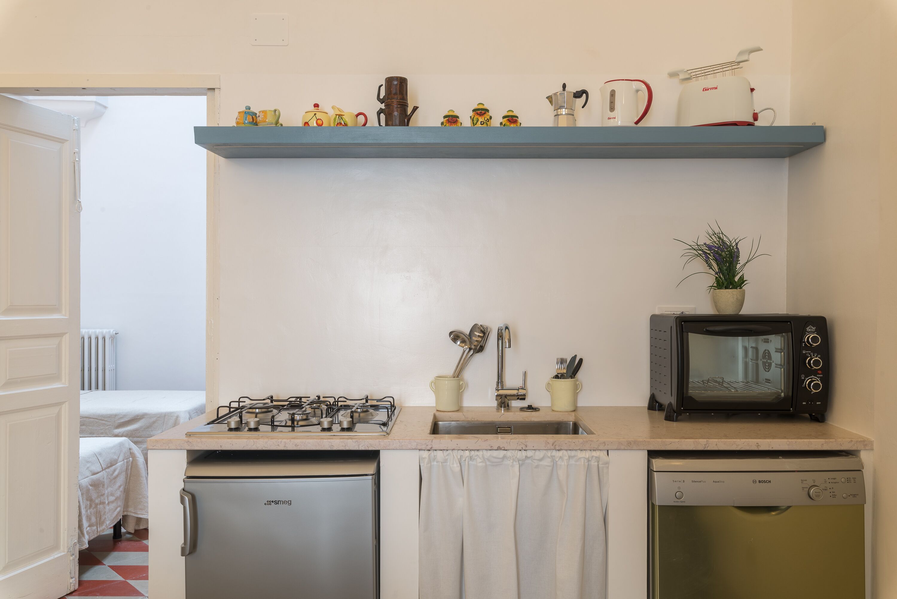 Une cuisine compacte équipée d’une cuisinière à gaz, d’un évier et d’un lave-vaisselle Bosch. Un petit four grille-pain est posé sur le plan de travail, et une étagère au-dessus contient divers ustensiles de cuisine.