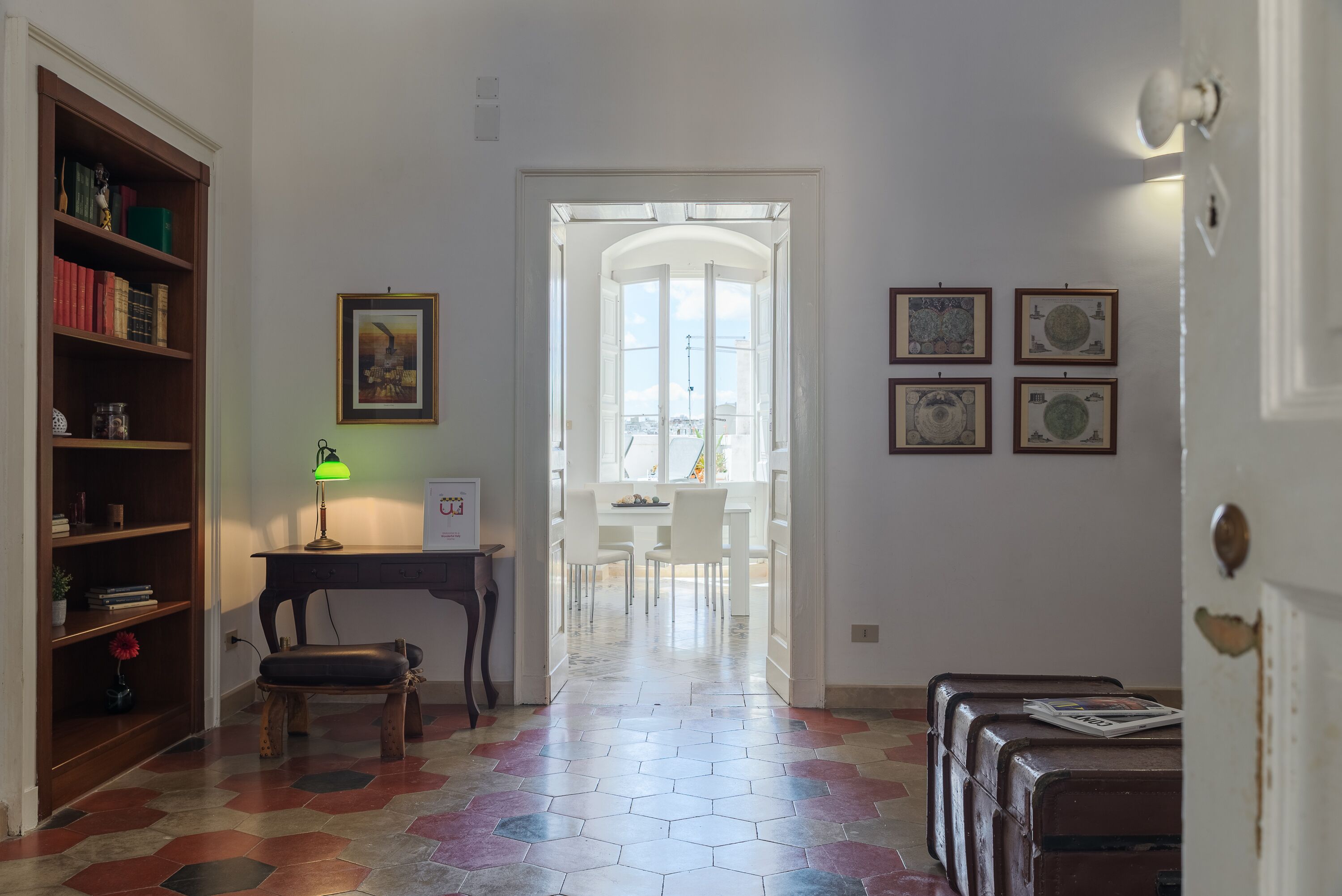 A hallway with a wooden bookshelf, a desk with a lamp, a dining area with chairs, and a view through an arched doorway.