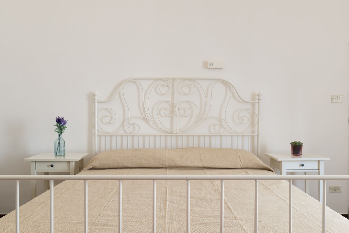 A bed with a white metal frame and a beige mattress. There is a small bedside table with a vase of flowers and a potted plant.