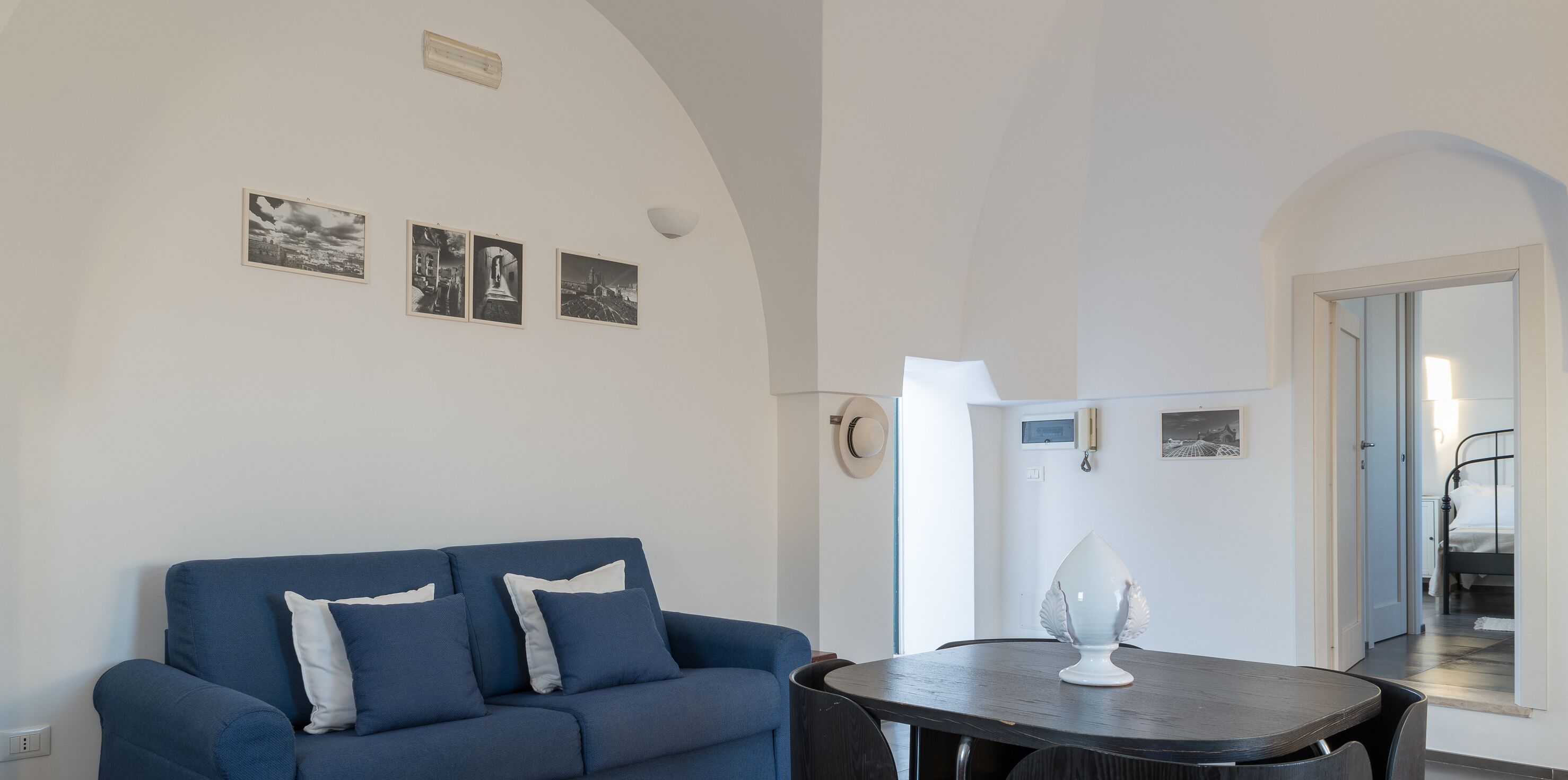 A modern living room with a blue sofa, a dining table with a decorative vase, and a hallway leading to a bedroom.