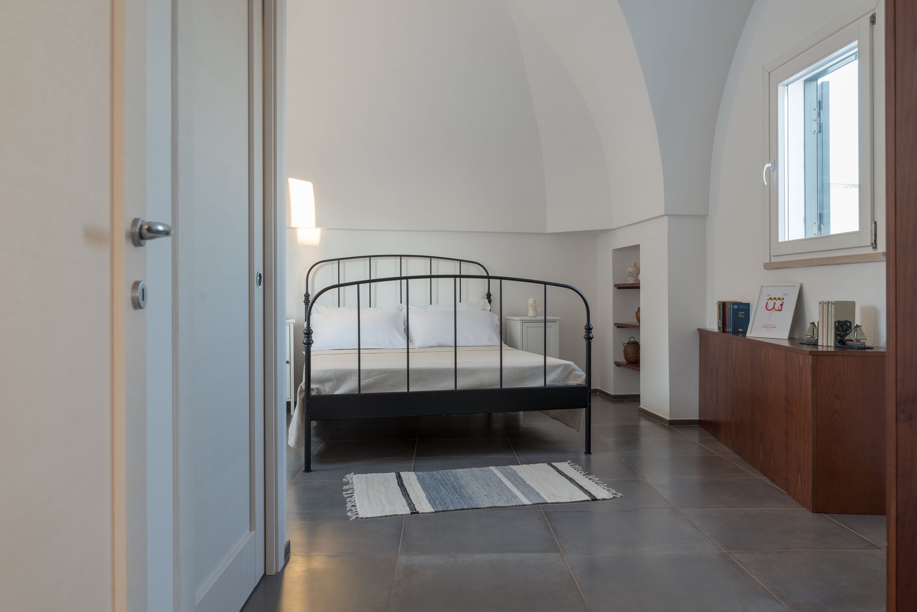 Un dormitorio con una cama, una mesita de noche, un escritorio de madera, una ventana y una puerta.