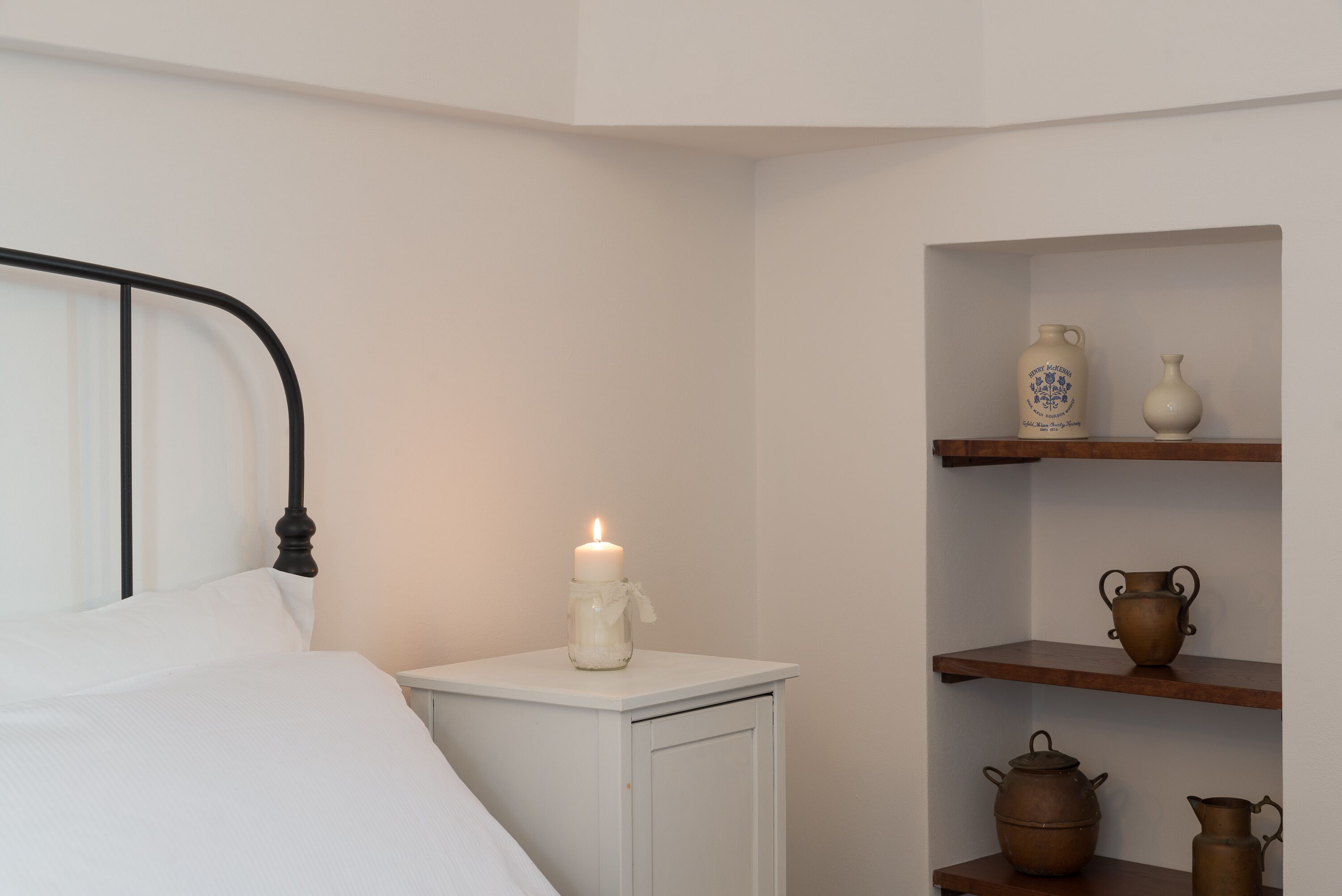 Un dormitorio con una cama blanca, una mesita de noche con una vela encendida y estantes con cerámica.
