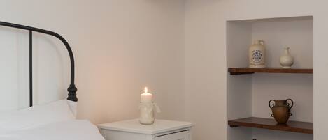 Un dormitorio con una cama blanca, una mesita de noche con una vela encendida y estantes con cerámica.