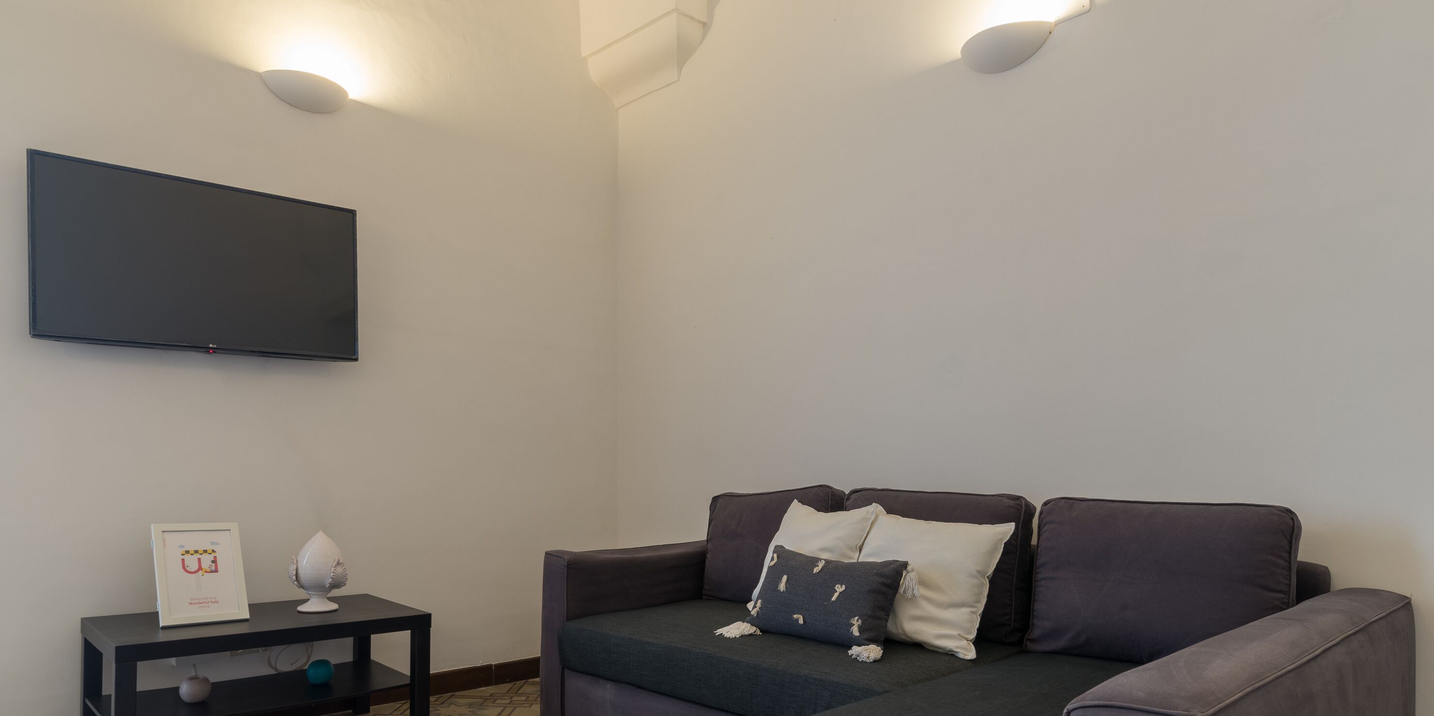 A modern living room with a flat-screen TV mounted on the wall, a dark-colored sofa with decorative pillows, and a small side table with a framed picture and a decorative item.