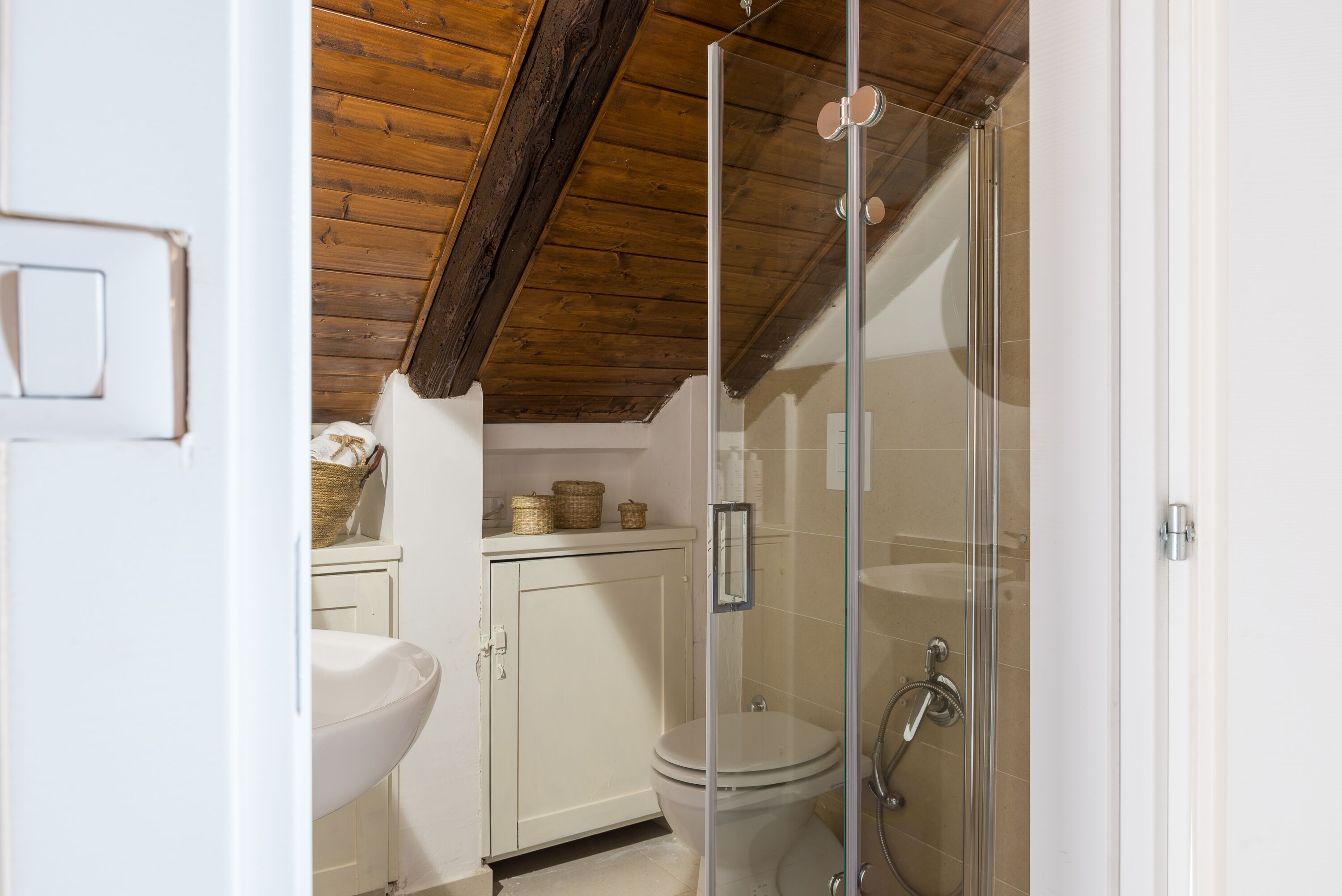 A bathroom with a white sink, toilet, and glass-enclosed shower. There is a wooden ceiling and a white cabinet with baskets.
