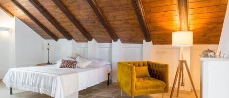 A bedroom with a wooden ceiling, a bed with white bedding, a mustard-colored armchair, a blue ottoman, and a floor lamp.