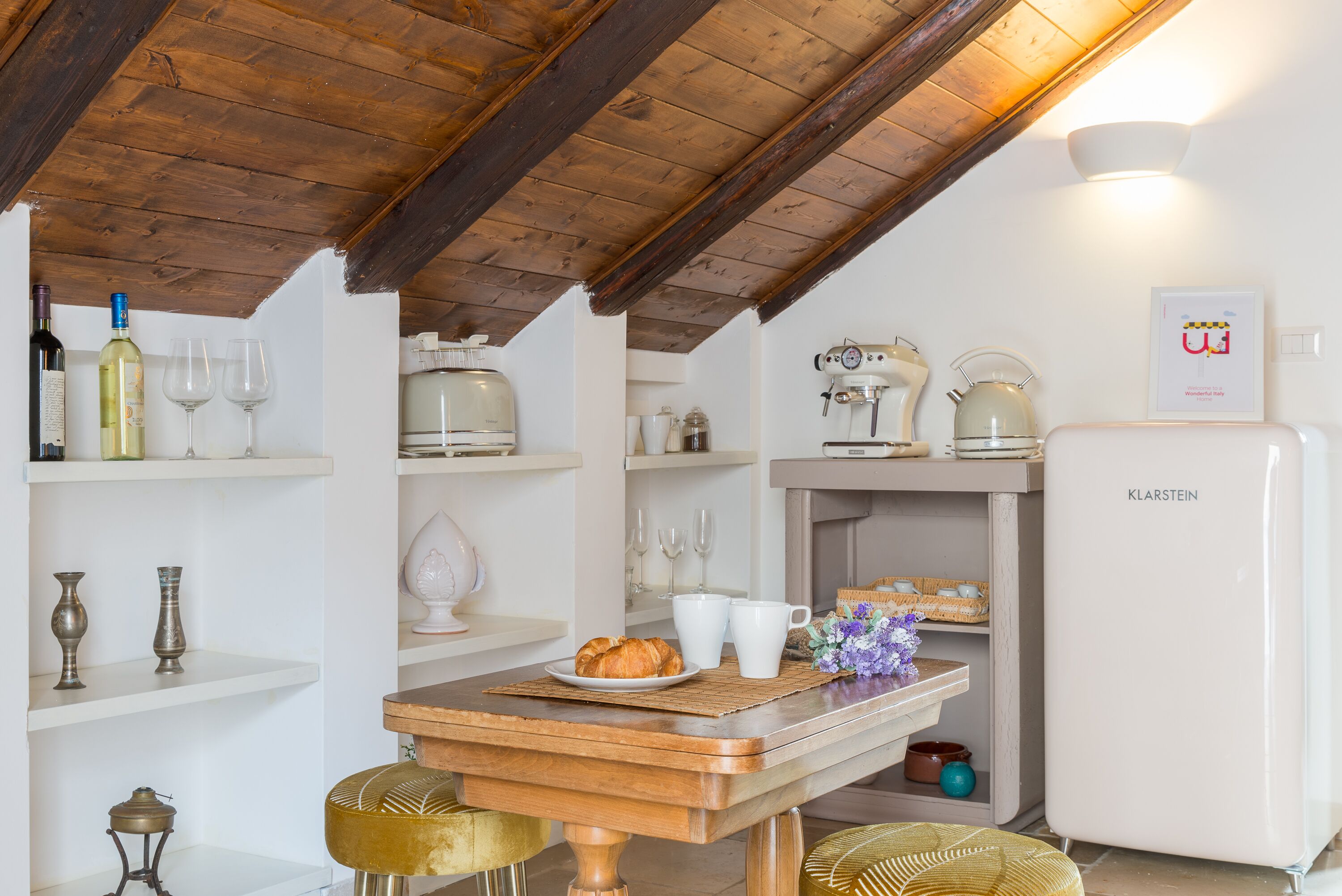 A kitchen with wooden beams, white cabinets, a K Klarstein refrigerator, and a table with a plate of croissants.