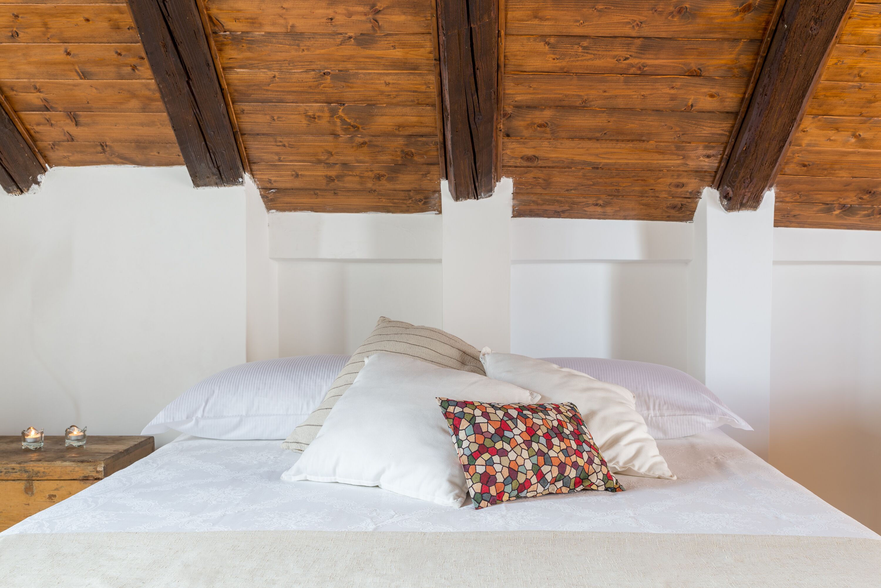 A bedroom with a wooden ceiling, a bed with white bedding, and two bedside tables with candles.