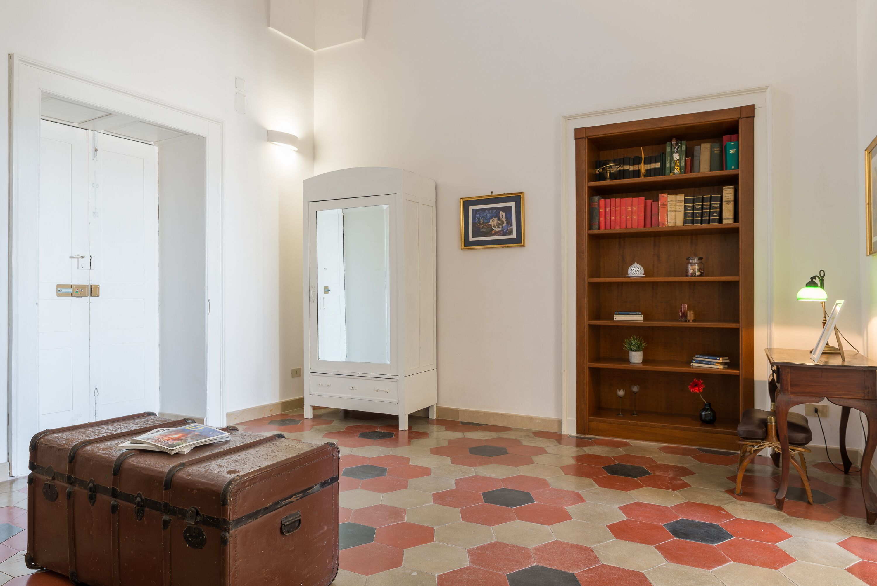 A room with a large wooden bookshelf, a white wardrobe, a brown suitcase, and a hexagonal patterned floor.