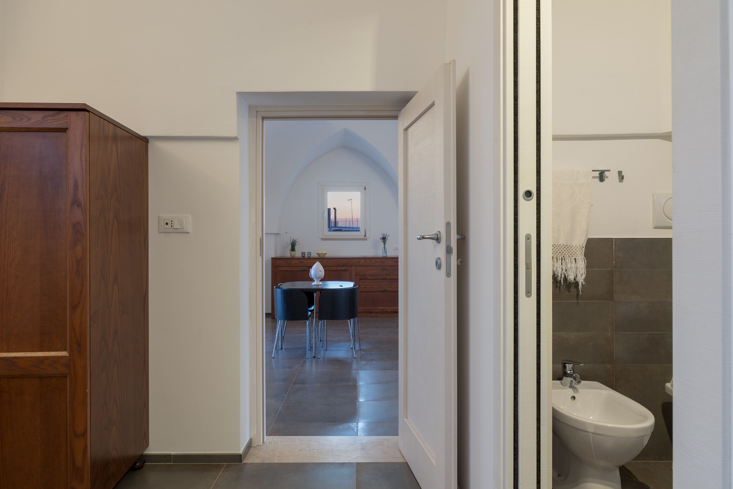 Um corredor que dá para uma sala de jantar com mesa e cadeiras, um armário de madeira e um banheiro com bidê.