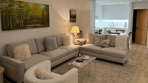 Un salon moderne avec un canapé d’angle, un fauteuil rond et une table basse. On peut voir un tableau au mur, une lampe et une vue sur la cuisine.