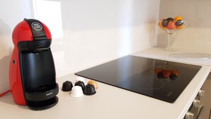 A red and black coffee maker with a glass carafe, a black induction cooktop, and a glass container with fruit on a white countertop. - City Center Apartment Project (Heraklion)