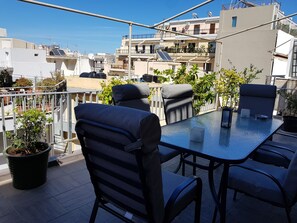 A rooftop terrace with a glass table and four chairs, surrounded by potted plants and overlooking a residential area.
