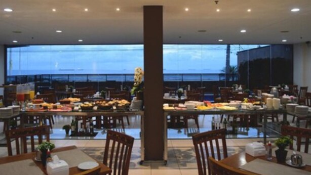 A spacious dining area with multiple tables and chairs, large windows offering a view of the sea, and a central pillar. - PraiaBella Hotel (São Luís)