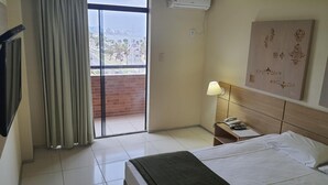 A hotel room with a bed, a nightstand, a lamp, a painting, and a view of palm trees through a sliding door. - PraiaBella Hotel (São Luís)