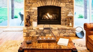 A stone fireplace with a wooden cart in front, a bookshelf above, and a view of the outdoors through large windows.