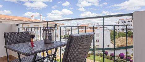 A balcony with wicker furniture, a glass of red wine, and a view of buildings and a swimming pool.