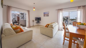 Un salon avec un canapé, une table à manger avec des chaises, un téléviseur et un balcon avec des rideaux.