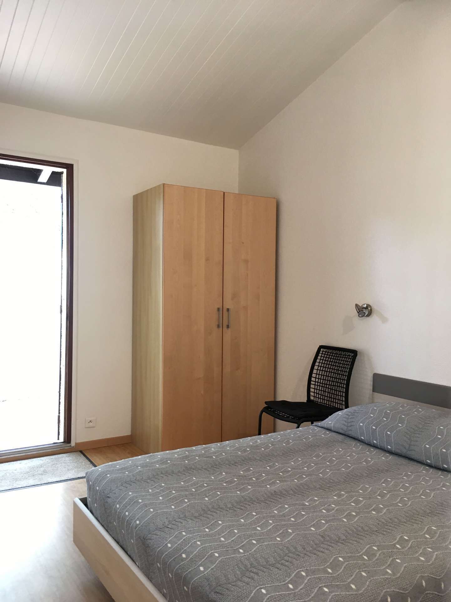 A bedroom with a bed, a wooden wardrobe, a chair, and a window.
