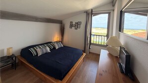A bedroom with a bed, a bedside table, a lamp, a TV, and a window with a view of the beach.