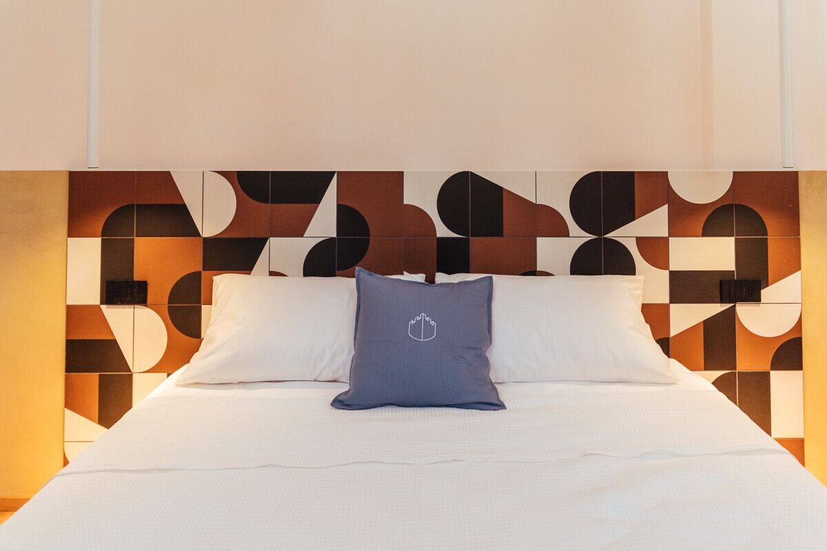 Quarto de hotel moderno com cabeceira estampada em padrão geométrico, cama bem arrumada com lençóis brancos e um único travesseiro azul com o logotipo de um brasão.