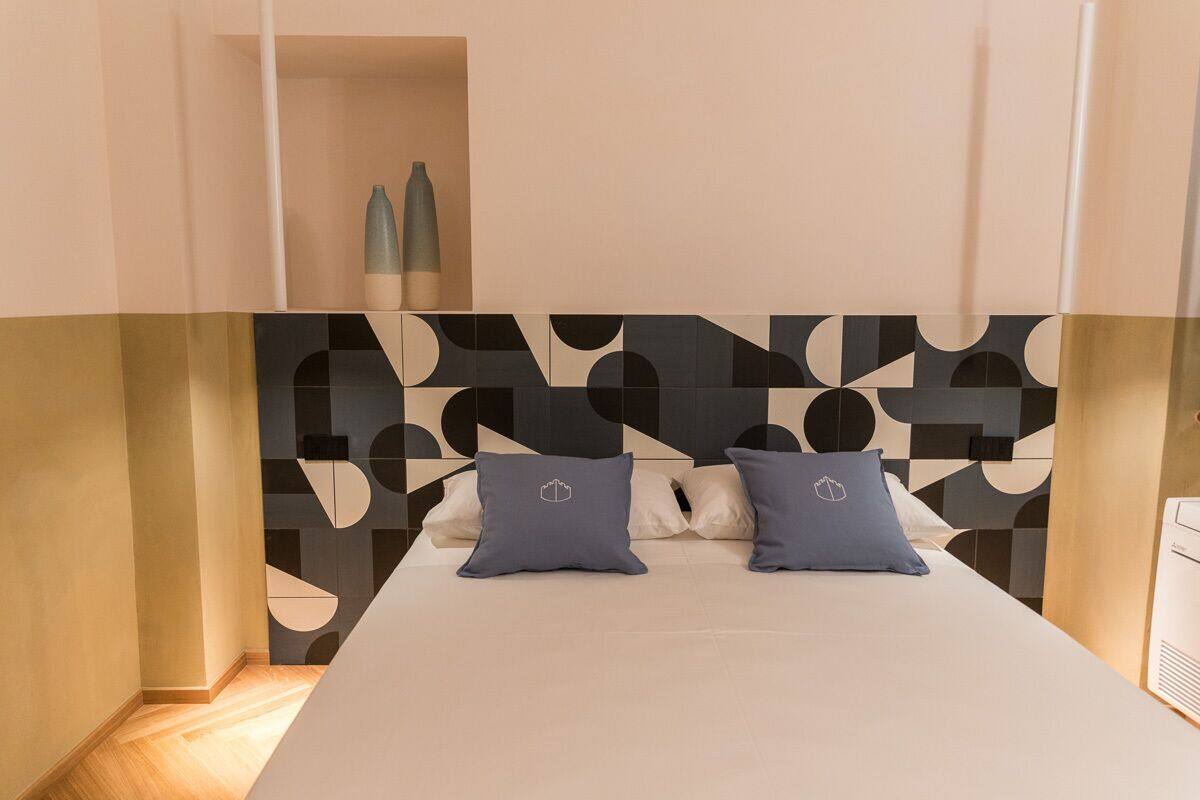 Quarto moderno com cabeceira estampada em padrão geométrico, uma cama grande com duas almofadas e uma mesa de cabeceira com abajur.