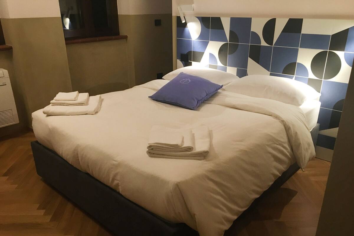 Uma cama bem arrumada com toalhas dobradas e um travesseiro azul, chão de madeira e parede com padrão geométrico.