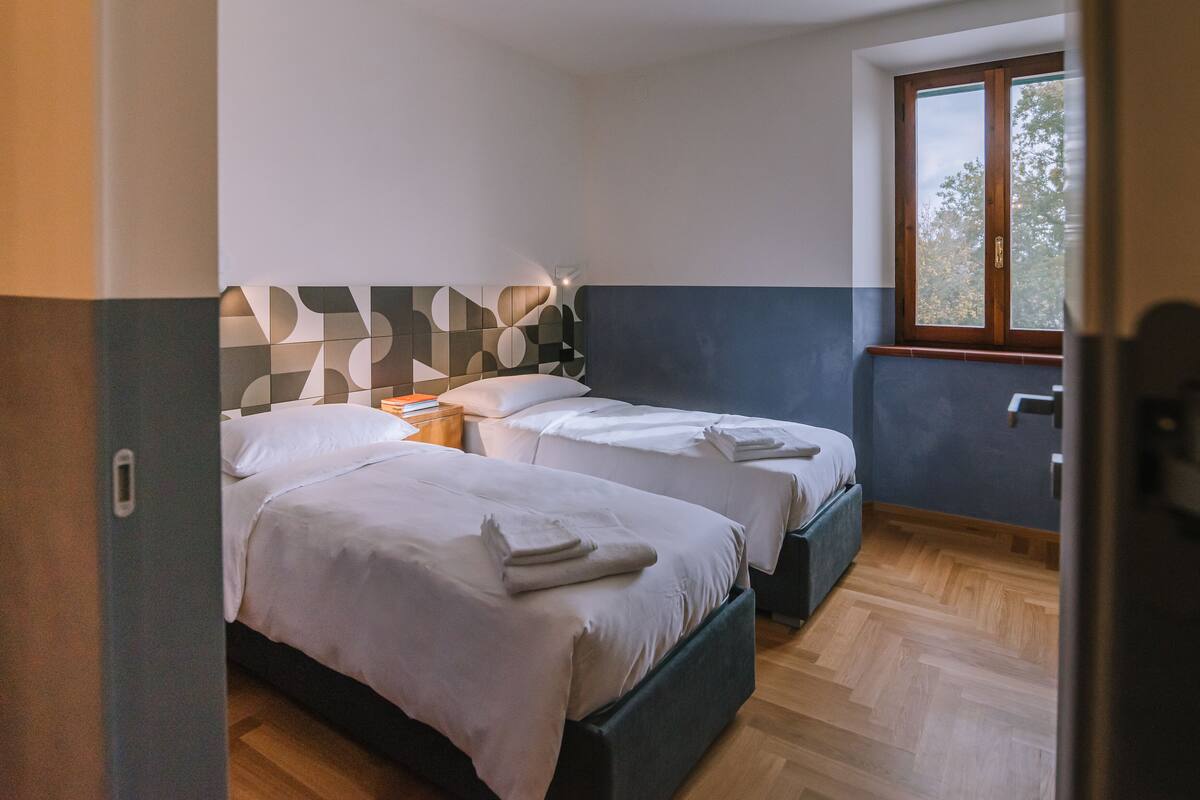 Quarto de hotel com duas camas, piso de madeira, uma janela e um design geométrico na parede.