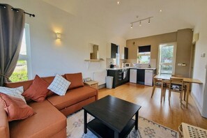 A compact living space with a sofa, a coffee table, a dining area with a table and chairs, a kitchenette with a microwave, and a window with curtains.