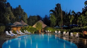 A pool area with lounge chairs and umbrellas, surrounded by tropical vegetation and thatched-roof structures. - The Rangers reserve Jim Corbett (Nainital)