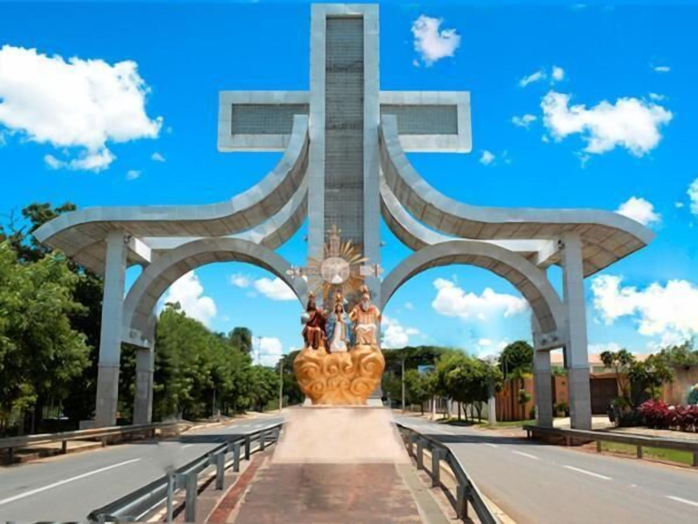 Um grande monumento em forma de cruz com uma estátua central, ladeada por duas grandes arcadas numa estrada larga com árvores e céu claro ao fundo.
