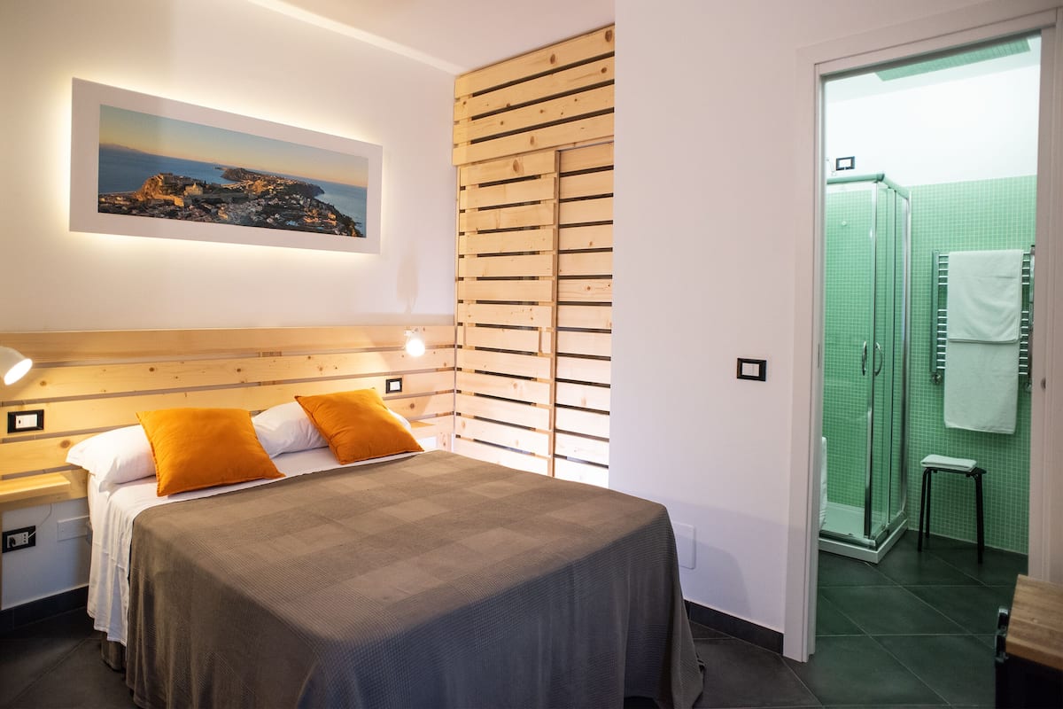 A bedroom with a bed, wooden headboard, a nightstand, a bedside table, a framed picture on the wall, and a view of the bathroom with a shower and a towel rack.