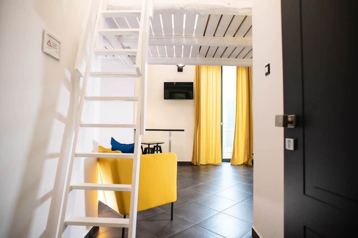 A modern interior with a white ladder, a yellow chair, a flat-screen TV, and a kitchen area with yellow curtains.