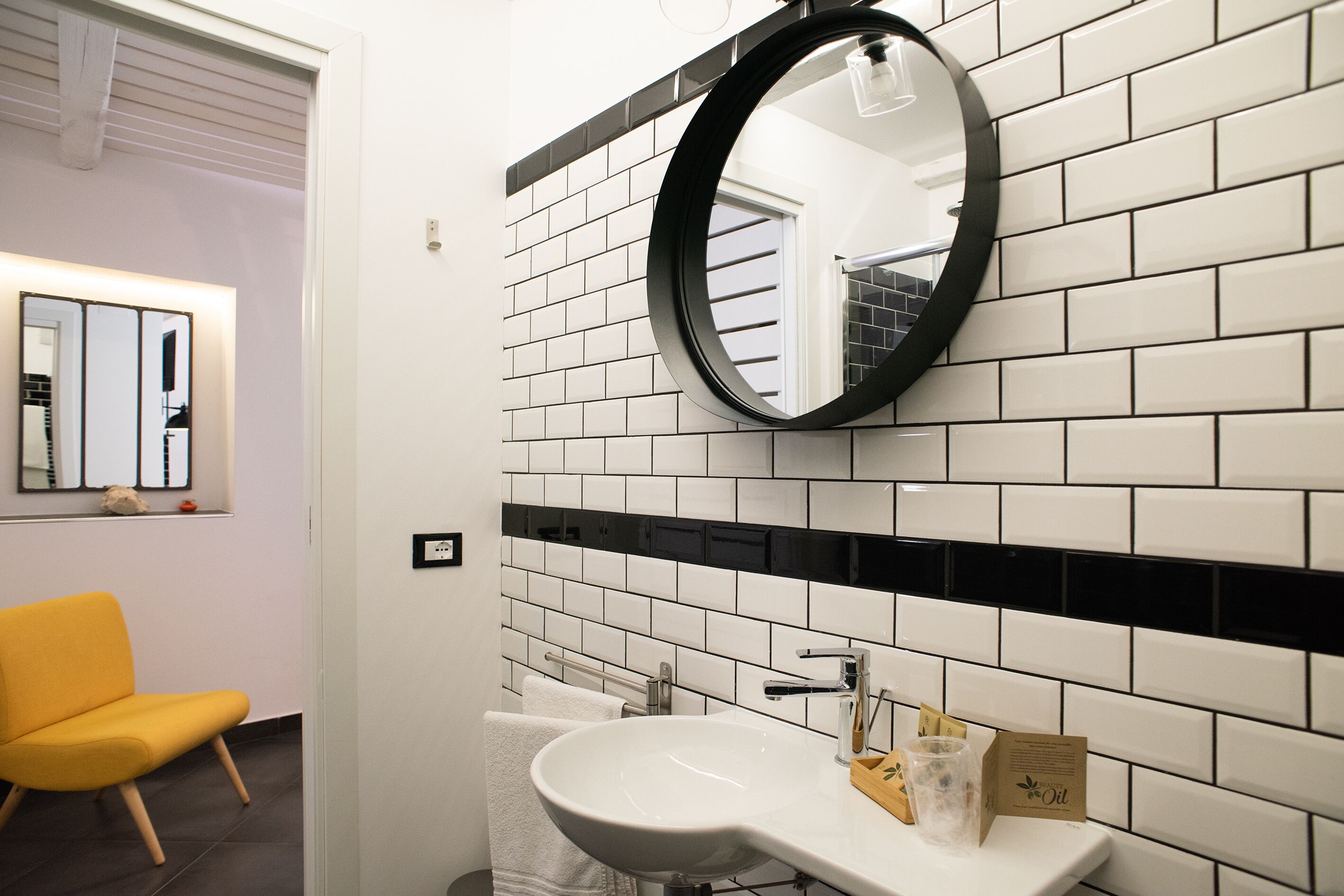 A bathroom with a white sink, black-tiled walls, a round mirror, and a yellow chair.