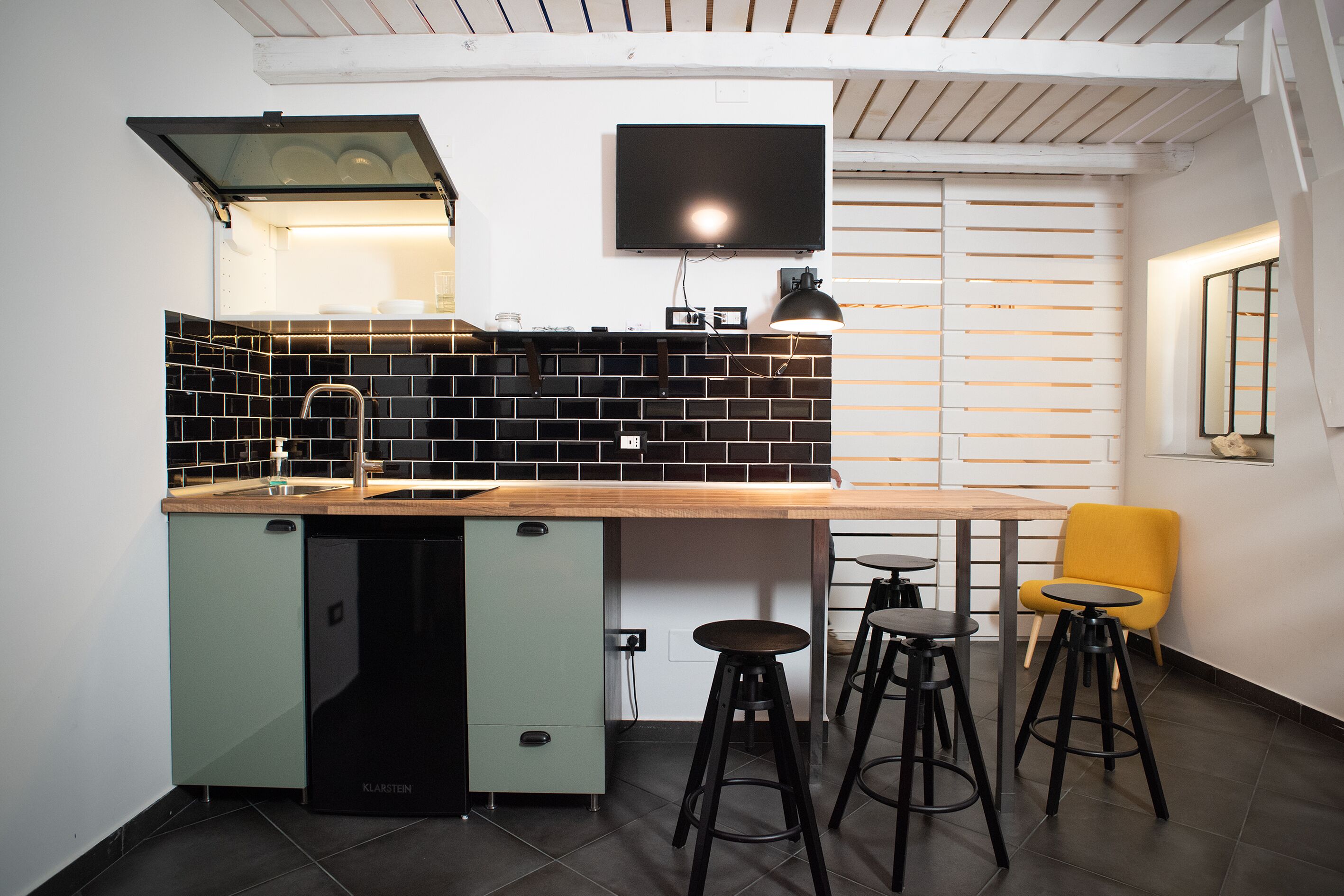 A compact kitchen with a built-in dishwasher, a wooden countertop, and a black tiled backsplash. There are two stools and a yellow chair near a window.