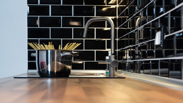 Une cuisine moderne avec une casserole en acier inoxydable remplie de spaghettis posée sur un plan de travail en bois, un robinet élégant et un dosseret de cuisine en carreaux noirs.