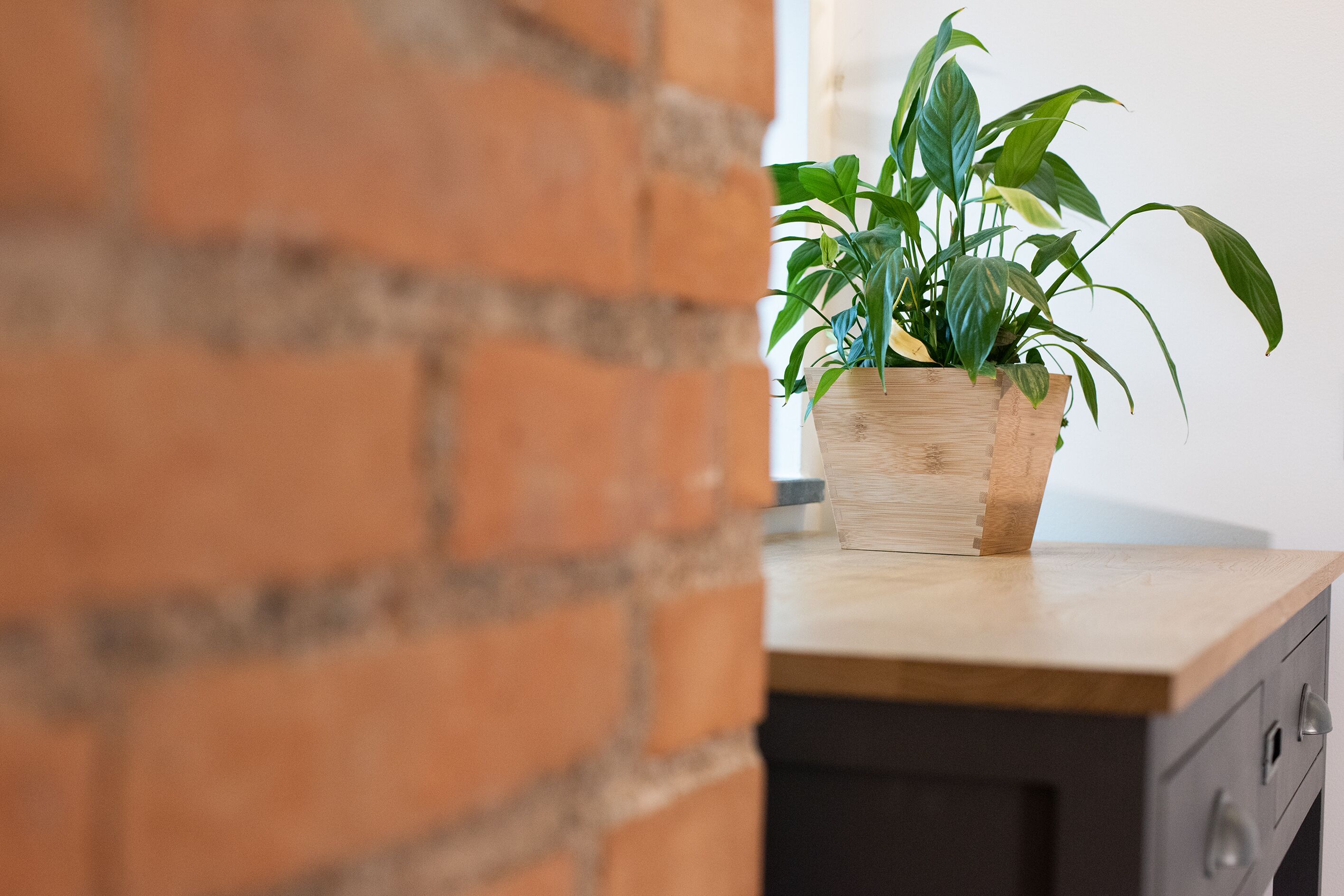 Um cachepot de madeira com plantas verdes sobre uma mesa de madeira, com um muro de tijolos ao fundo.