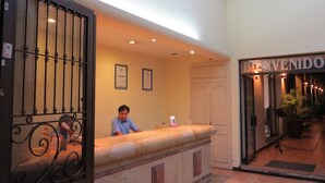 Un mostrador de recepción de hotel con un recepcionista, un cartel de 'BIENVENIDO' y certificados enmarcados en la pared.