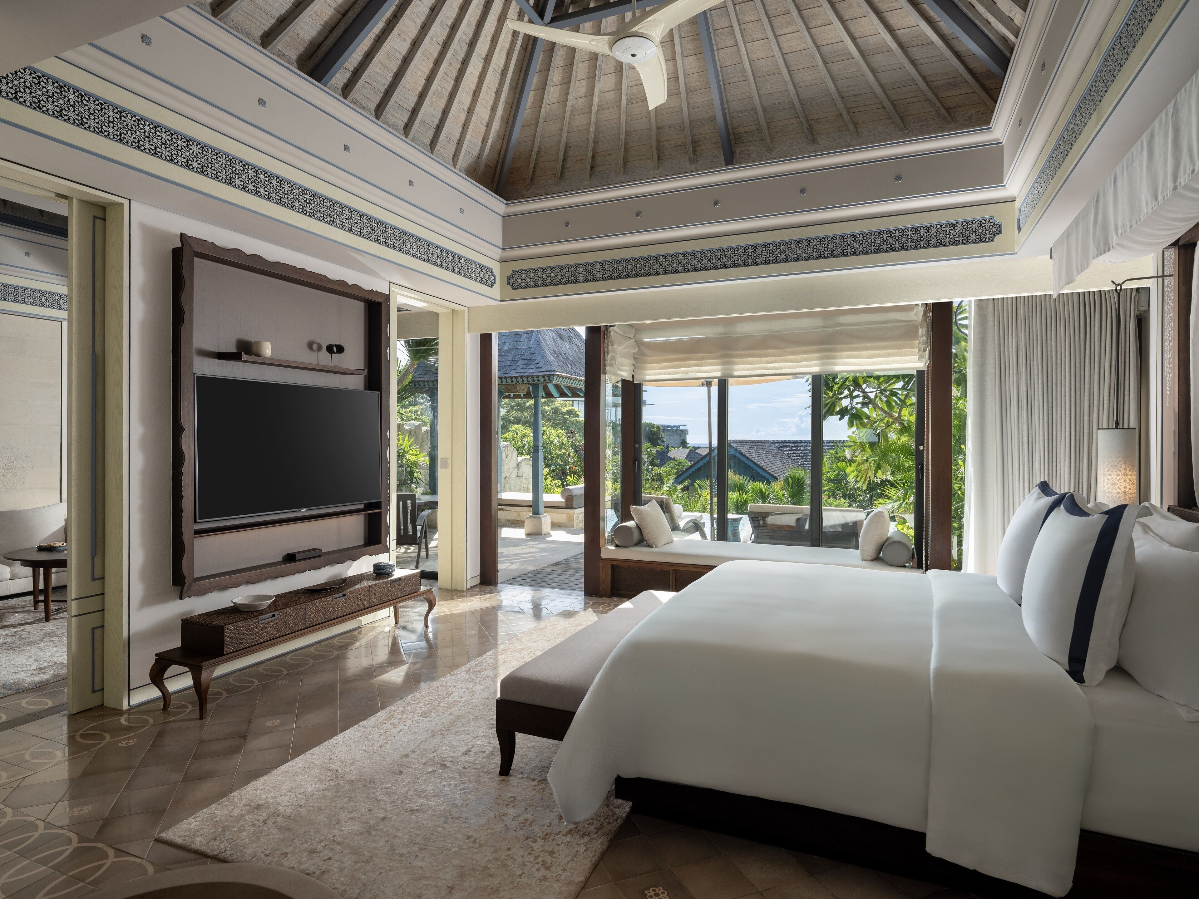 A spacious bedroom with a large bed, a flat-screen TV, and a view of a tropical garden through large windows.