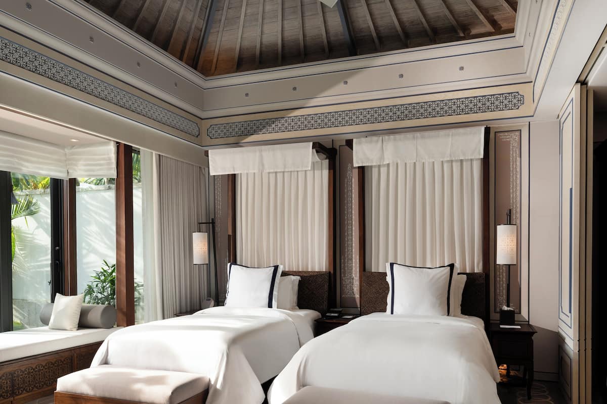 A hotel room with two beds, a large window with curtains, a wooden ceiling, and a nightstand with a lamp.