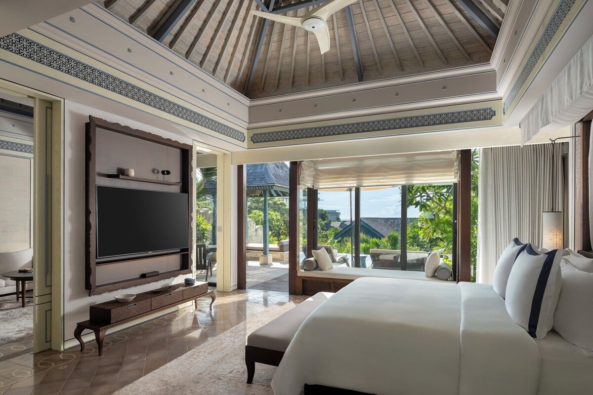 A spacious bedroom with a large bed, a flat-screen TV, and a view of a lush garden.