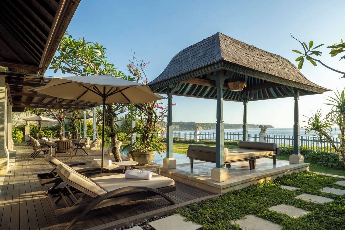 A covered outdoor seating area with lounge chairs and umbrellas, a wooden deck, and a view of the sea.