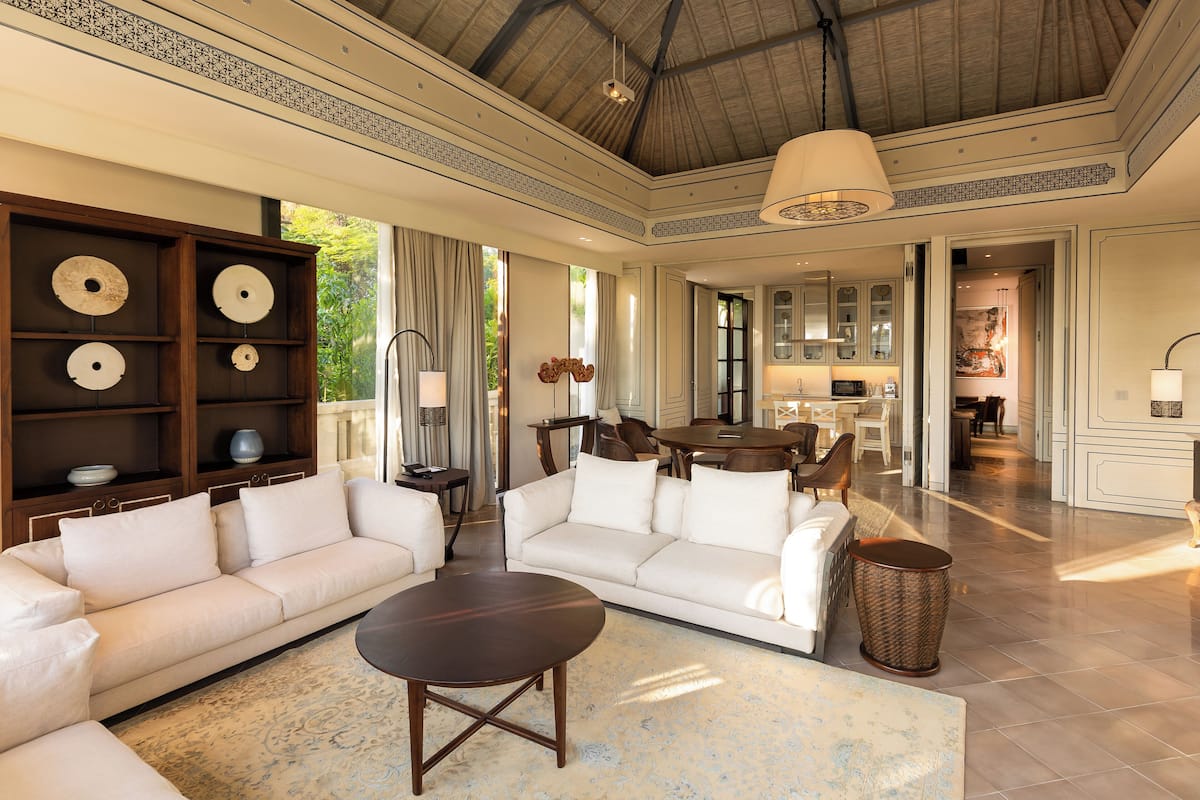 A spacious living area with a large sofa, a coffee table, and a dining table. There is a kitchen in the background with cabinets and a sink.