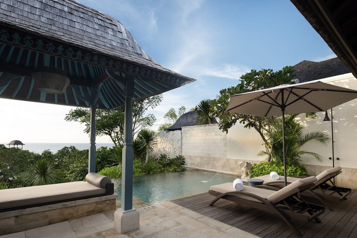 A poolside area with lounge chairs and umbrellas, a stone wall, and a view of the sea.