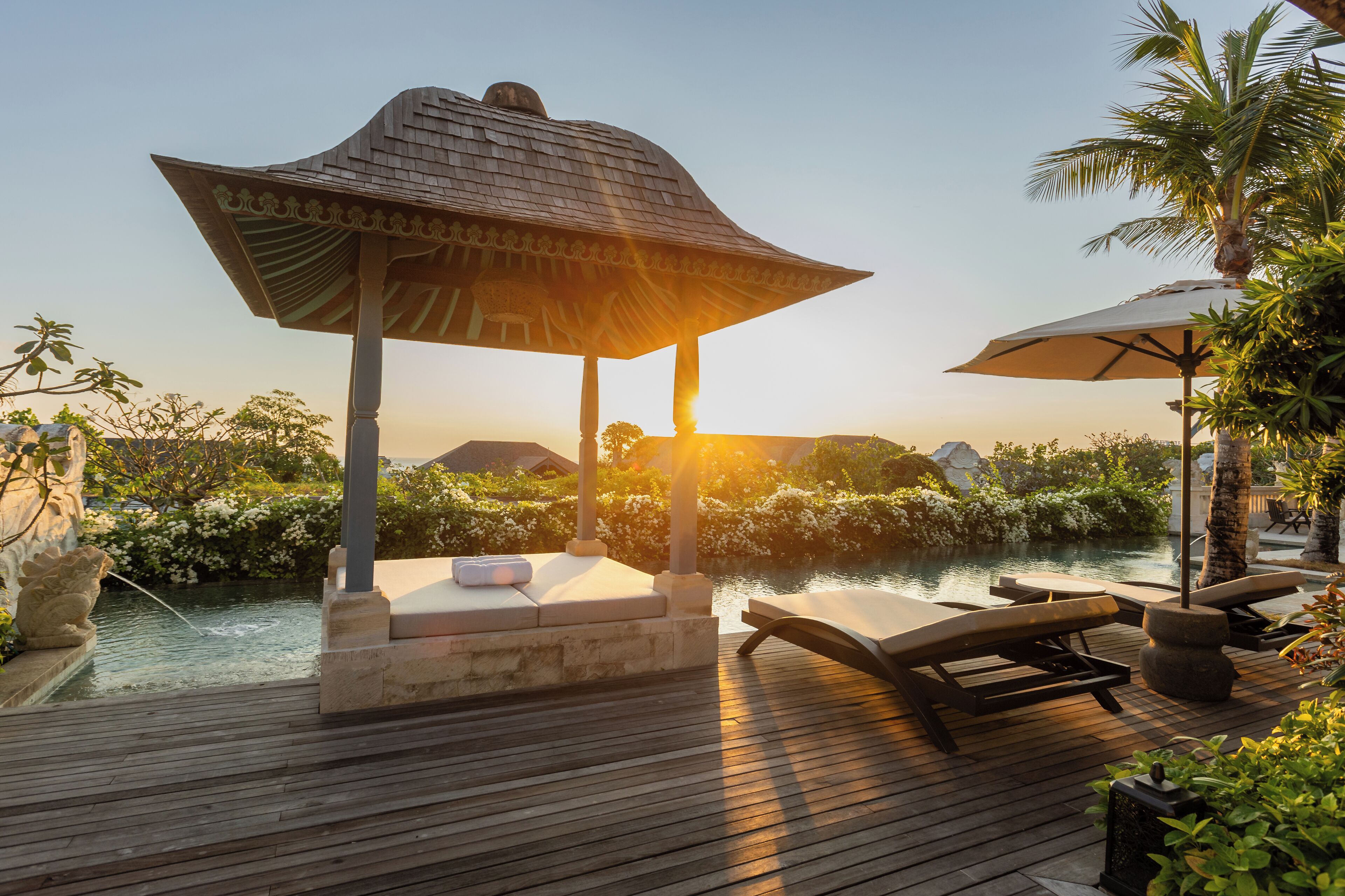 A wooden deck with lounge chairs and a stone fountain, overlooking a pool and lush greenery.