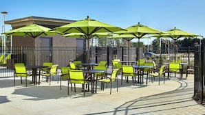 Un espace extérieur aménagé en terrasse, avec des tables et des chaises, des parasols verts et un bâtiment en arrière-plan.