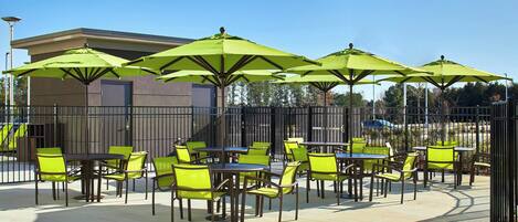 An outdoor patio area with tables and chairs, green umbrellas, and a building in the background.