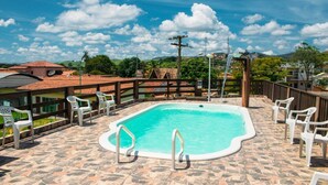 A swimming pool with white chairs and a stone-tiled deck, surrounded by a wooden fence and buildings in the background. - Pousada Campo Alegre (Itatiaia)