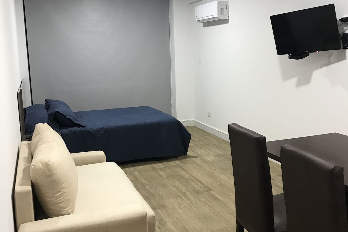 A compact room with a bed, a sofa, a table, and a TV mounted on the wall.